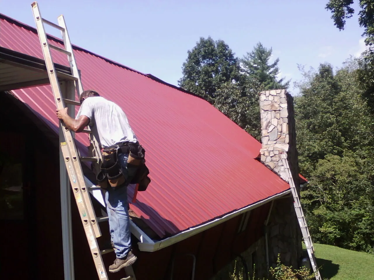Roofer on a ladder beside a red metal roof and stone chimney — Soffit & Fascia Repair by Roofing Brothers in Crescent City